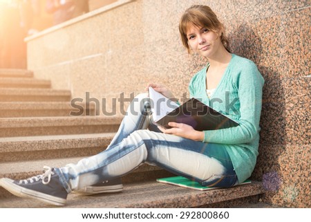 Portrait of a beautiful teenage student girl sitting on stairs in park with copy book and studing. Sunny summer day. Modern and casual lifestyle. Woman with note pad.