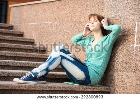 Young woman listening music in Headphones. Beautiful young female student.  Woman sitting on stairs in city park.