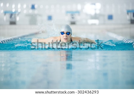 Young woman in goggles and cap swimming butterfly stroke style in the blue water indoor race pool