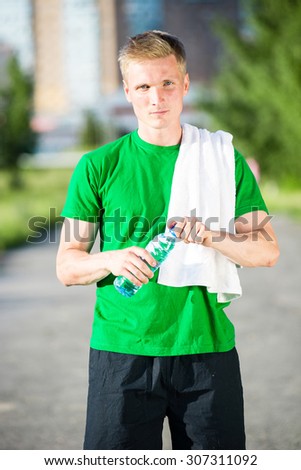 Tired man with white towel drinking water from a plastic bottle after fitness time and exercising in city street park at beautiful summer day. Sporty model caucasian ethnicity training outdoor.