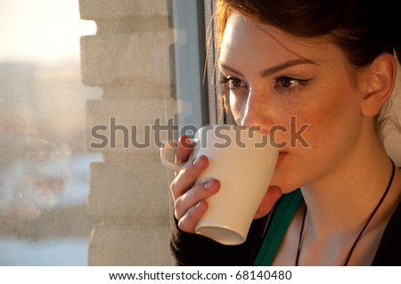 Beautiful young woman in casual wear dreaming with cup of hot coffee over window