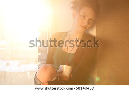 Beautiful young woman in casual wear dreaming with cup of hot coffee over window. Sun beam.