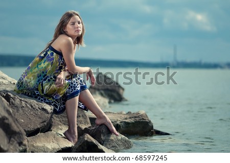 Beautiful sad woman in dress sitting on rock over sea at summer storm day