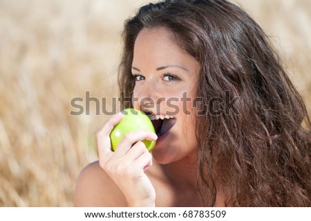 Beautiful woman with perfect hair and skin posing in wheat field and eating green apple. Summer picnic.