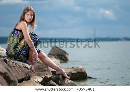 Beautiful sad woman in dress sitting on rock over sea at summer storm day