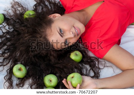 Beautiful woman in red dress with perfect hair and skin lying in wheat field with green apple. Picnic.