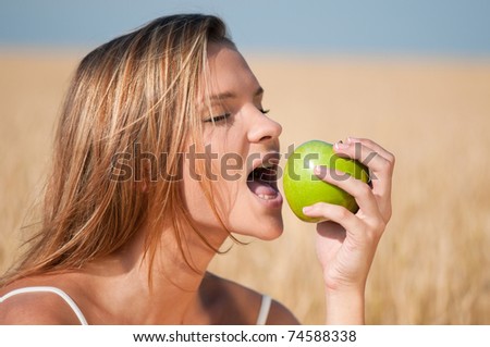 Beautiful woman with perfect hair and skin posing in wheat field and eating green apple. Summer picnic.