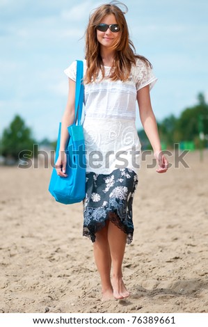 Beautiful young woman in dress and sunglasses walking on sand beach with bag on sunny summer day