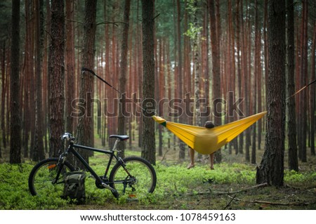 Bike trip to the forest. Bicycle and green military backpack in the focus, man relaxing in the hammock hanging among the red pine trees in the background. Selective focus. Travel. Bike packing concept