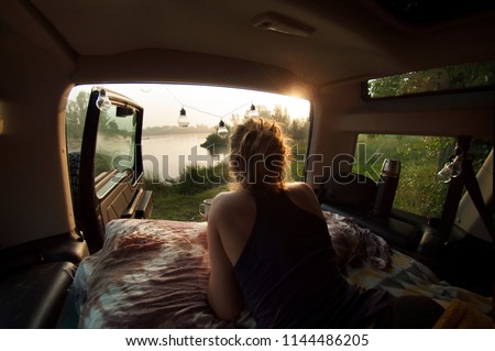 Woman camping in the camper car and drinking morning coffee. Beautiful calm lake in the background. Sunrise mild natural light.  Wild camping concept. Horizontal image.