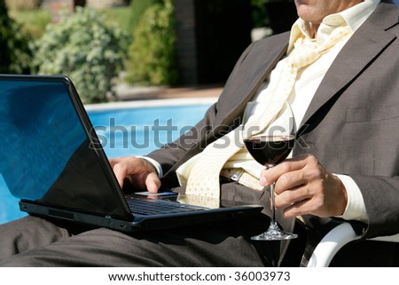Businessman sitting next to the swimming pool with laptop and glass of red wine