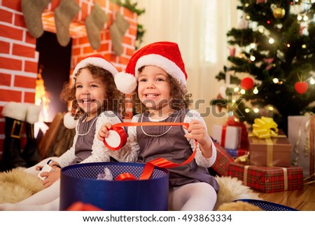 Little girls twins at Christmas sitting on the floor in Santa hats smiling and laughing in a room decorated in a happy new year.