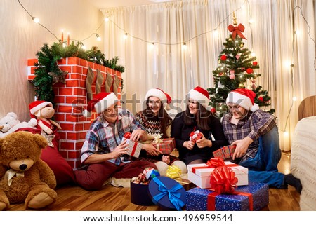 Group of friends at Christmas, New Year sitting on the floor with gifts near tree and fireplace.