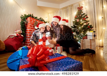 In a group of friends in christmas hats on the floor in the room to open presents with the Christmas tree. Magic light out of the box with a gift in a happy New Year.