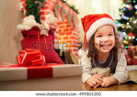 The little girl in Christmas hat of Santa Claus lying on the floor in the room with the decor, fireplace, smiling in the New Year.