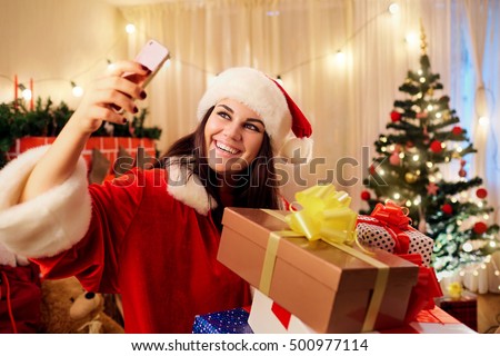 Beautiful girl in the hat of Santa Claus with gifts in his hands makes slefi telephone in the room with the decor,  tree, fireplace, gift boxes at Christmas, Happy New Year.