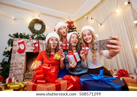 Beautiful girls, friends in caps of Santa Claus doing selfie mobile phone, laughing, smiling in Christmas room decoration new year.