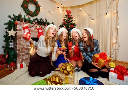 Friends sing karaoke at Christmas, New Year. Four beautiful girls in caps of Santa Claus singing with a microphone in the room with the decor.