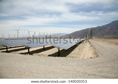 Solar Panels AKA- Photovoltaic Cells in a SOLAR FARM with Wind Turbines in the background collect and produce Electricity from Natural Renewable Resources of the Sun and Wind producing GREEN ENERGY