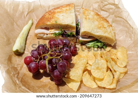 A picnic lunch with a Turkey and Cheese Sandwich on Cheese Bread, Chips, Red Grapes and a Dill Pickle Slice wrapped in deli paper, isolated on white. Deli Sandwiches are a favorite for picnics