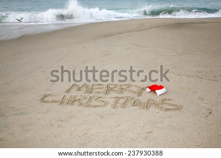 The words Merry Christmas written in the sand with a Red Christmas Santa Claus hat next to the word Merry with the Ocean and Seagulls in the background. Christmas is the time many go to the beach