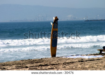 a lone surf board sits in the sand with the blue ocean and white waves in the background waiting for its owner to come back to go surfing