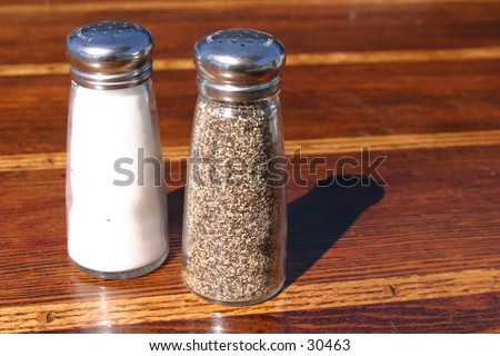 salt and pepper shakers sit upon a wooden table outside in the midday sun casting a shadow and glistening stainless steele tops
