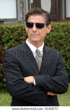a secret agent wears dark sunglasses and a dark suit while he looks around to protect his employer while at a high profile event