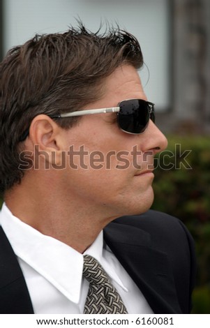 a secret agent wears dark sunglasses and a dark suit while he looks around to protect his employer while at a high profile event