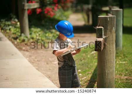 a young boy plays contractor while wearing a blue hard hat and taking notes on a clip board