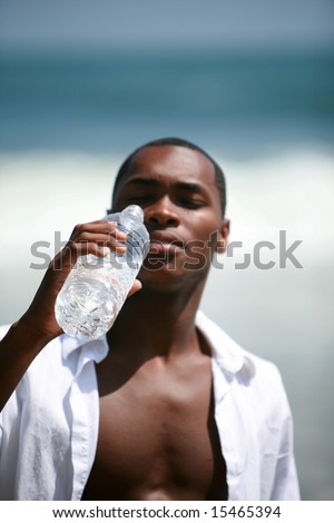 a handsome african american male model drinks fresh cold water from a bottle at the beach on a beautiful summer day