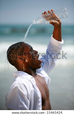 a handsome african american male model pours fresh cold water from a bottle on his head to cool down at the beach on a beautiful summer day