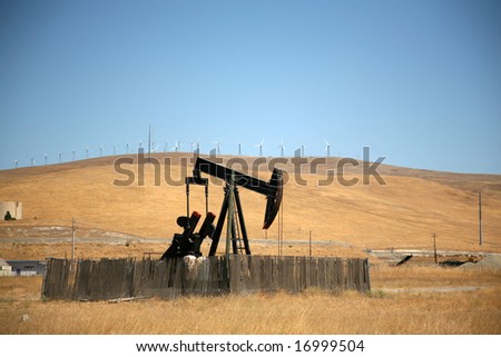 an oil pump pumps oil as Wind Mill Turbines Churn in the background representing New and Old Technology in the Energy Markets