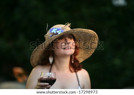 an attractive woman in a pretty and fun hat enjoys a glass of red wine by a pool one evening
