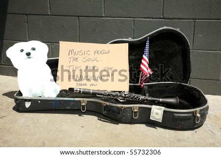 an empty guitar case with a sign and a cardboard cut out of a bichon frise dog asking for tips to play music