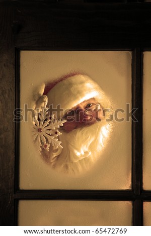 Santa Claus looks out through the snow and fog on his workshop window in the north pole to see outside check the weather while he holds up a large snow flake on December 24th, Christmas Eve