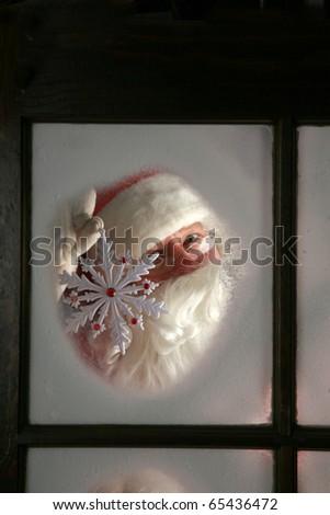 Santa Claus looks out through the snow and fog on his workshop window in the north pole to see outside check the weather while he holds up a large snow flake  on December 24th, Christmas Eve