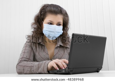 The young woman at office in medical mask works with the laptop