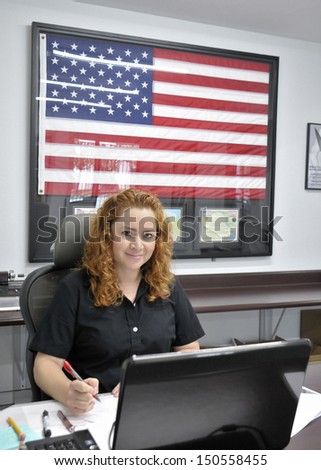 Pretty Hispanic Woman Receptionist at desk with a computer and American Flag in the background