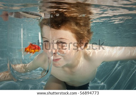 boy making funny face swimming underwater in swimming pool looking at goldfish in glass bowl