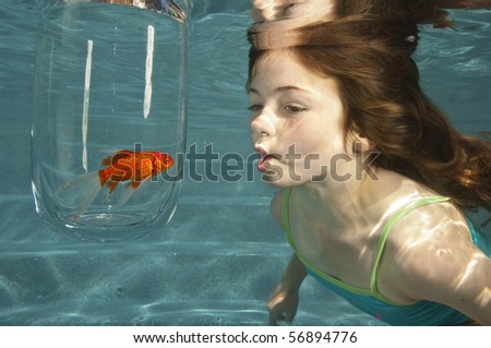 girl making funny fish face swimming underwater in swimming pool looking at goldfish in glass bowl