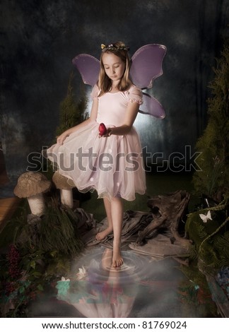 little girl dressed as fairy dipping toe in pond and holding red bird