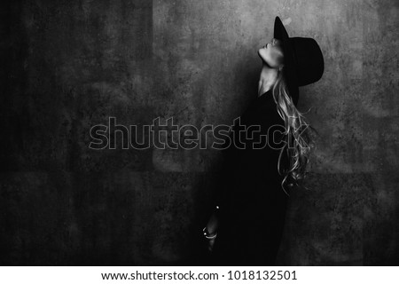 Beautiful young mysterious blond girl in black hat and black jacket on gray background. Eyes are covered with a hat. Manicure - long red nails, nail polish. Fashion, beauty.