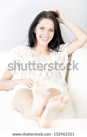 Beautiful young woman with long dark hair lying on her back on a white sofa at home with her feet up