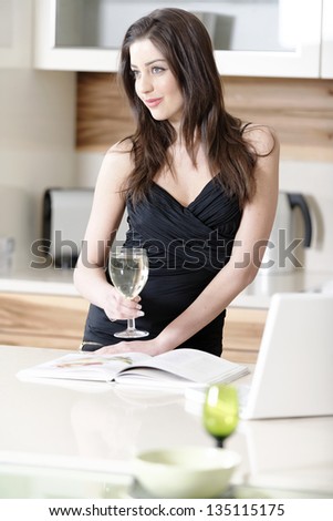 Attractive young woman using a laptop while enjoying a glass of wine in the kitchen