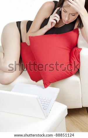 Young woman at home on the sofa, expressing worry on the phone while using a laptop.