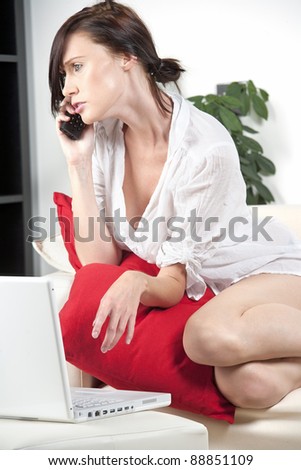 Young woman at home on the sofa, expressing worry on the phone while using a laptop.