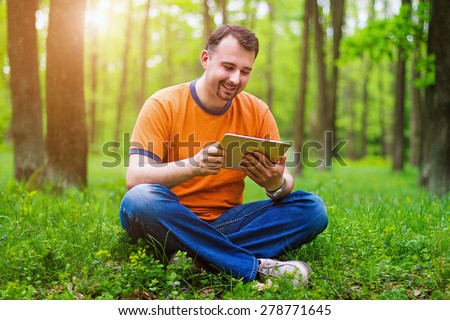 Handsome middle-age man in orange t-shirt sitting on the grass in the park and holding a tablet. He is smiling, sun is shining on the background