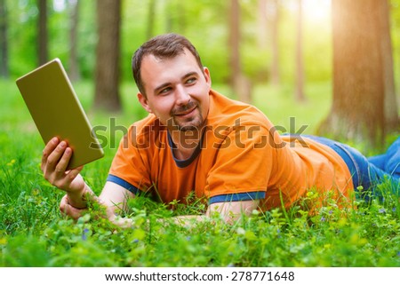 Handsome middle-age man in orange t-shirt lying on the grass in the park and holding a tablet. He is smiling, sun is shining on the background