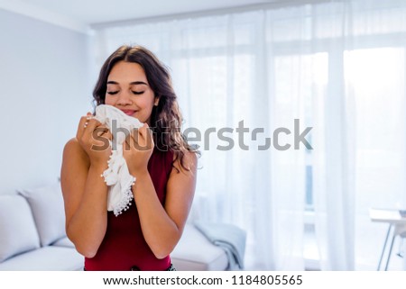 Beautiful young woman is smelling clean clothes and smiling while doing laundry at home. Woman Smelling Clean Clothes. Satisfied brunette lady enjoying nice smell of clean washed clothes.